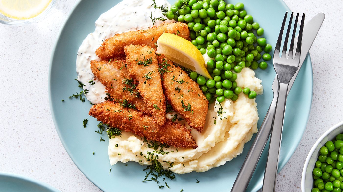 Cinq bouchées de poisson pané dorées servies avec une purée de pommes de terre, des petits pois et un dip dans une assiette bleue.