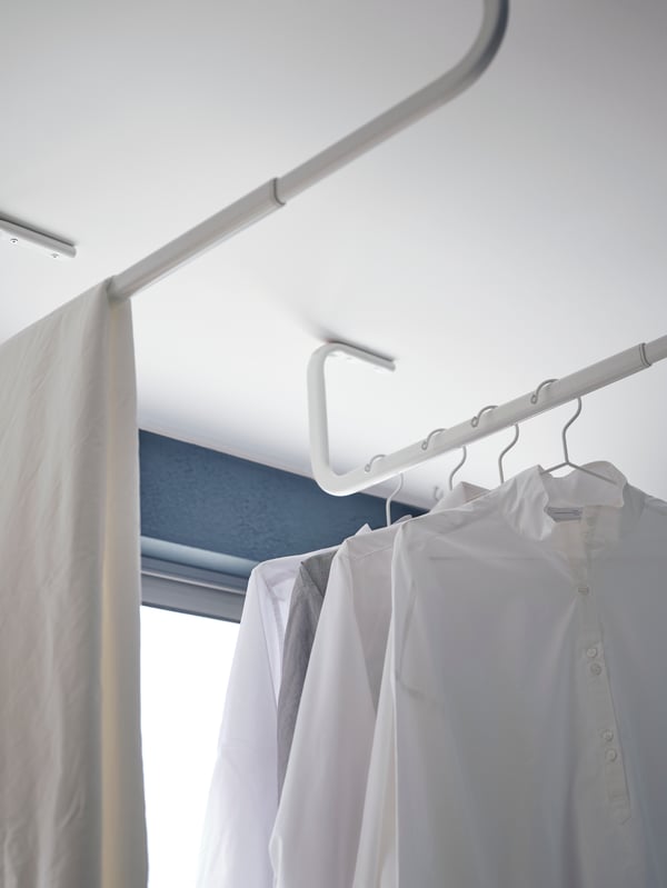 Dos colgadores de ropa MULIG blancos instalados al techo, cerca de una ventana y con camisas colgadas. 