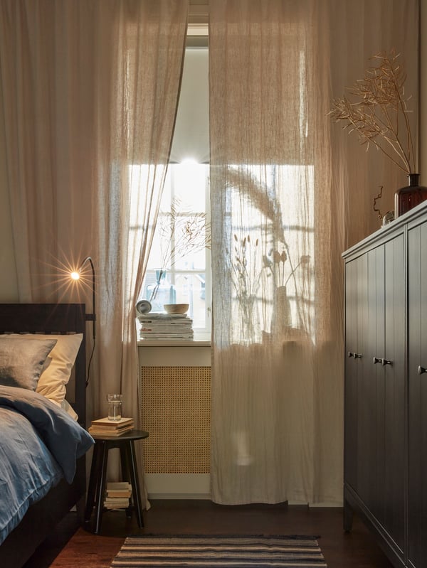 Gemütliches Schlafzimmer mit sanftem Tageslicht, das durch die transparenten DYTÅG Vorhänge fällt. Es gibt ein ordentlich gemachtes IDANÄS Bett, einen kleinen Beistelltisch mit Büchern und einen dunklen IDANÄS Kleiderschrank, die eine ruhige Atmosphäre schaffen.