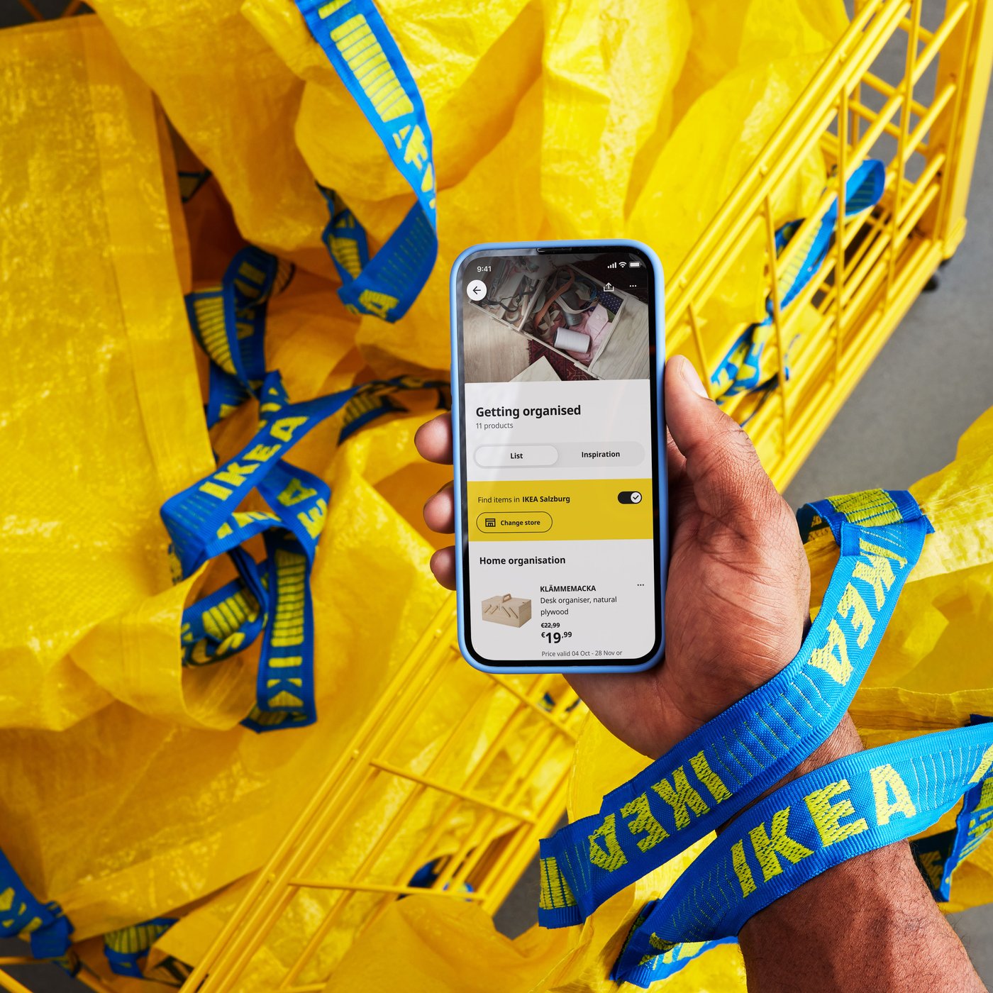 A hand holds a smartphone displaying the IKEA app, surrounded by yellow shopping bags used in-store.