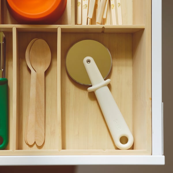An UPPDATERA cutlery tray in light bamboo with an UPPFYLLD pizza cutter, a green potato peeler and three wooden spoons.