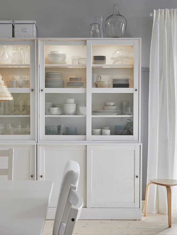 HAVSTA storage combination with white glass doors with items on the shelves and a lamp, decorations, and plants on top.