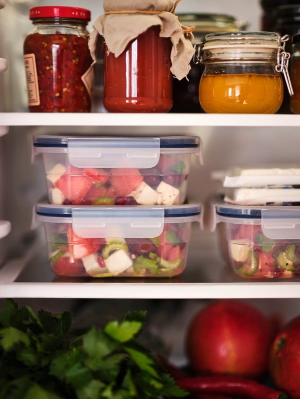 Shelves inside an open fridge, with bottled preserves, salad in square 750ml IKEA 365+ food containers and fresh vegetables.