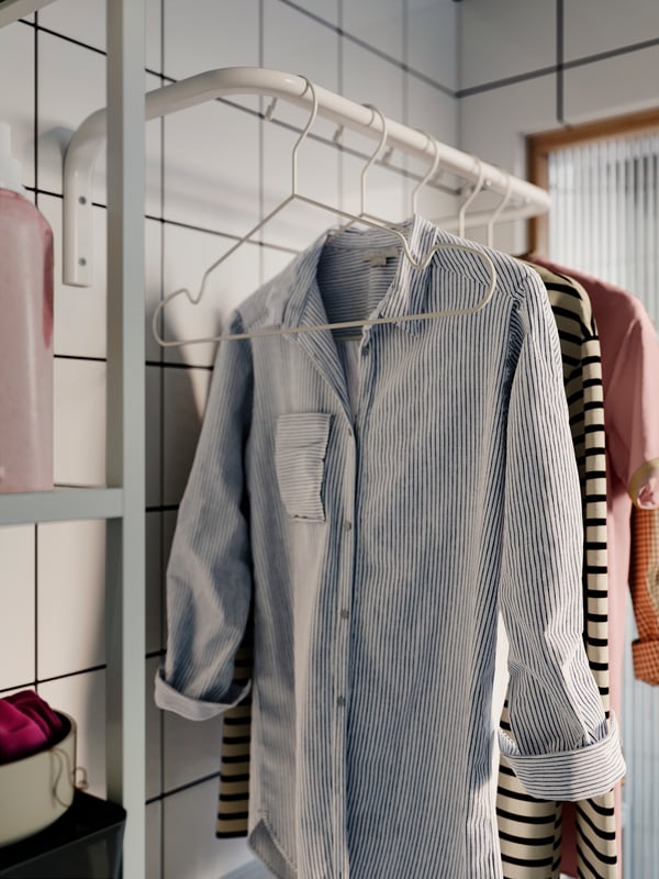 Drie gestreepte shirts hangen op een rij op witte kleerhangers aan een witte, wandgemonteerde MULIG kledingroede in een witgetegelde badkamer.