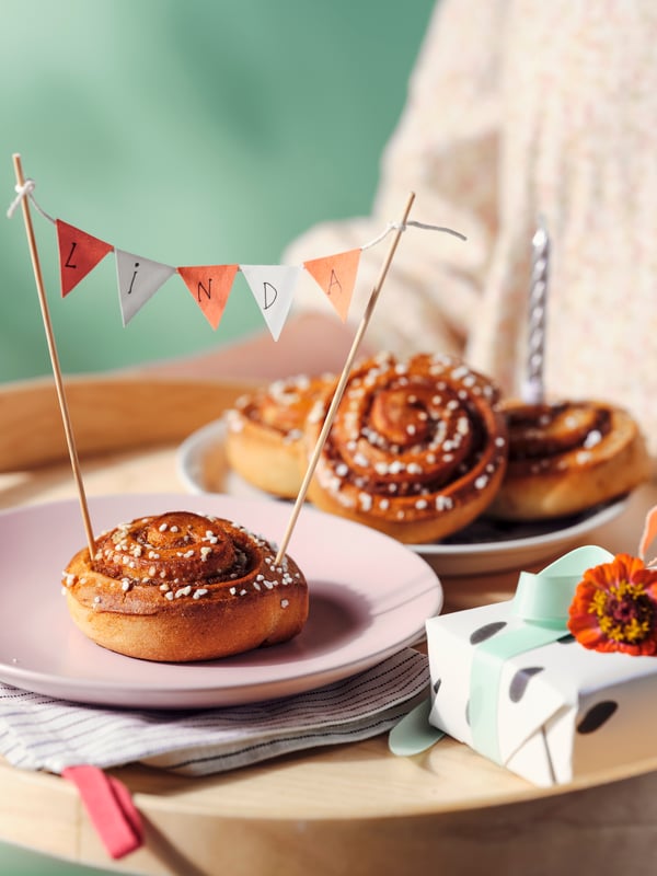 A person holding a wooden tray of KAFFEREP cinnamon buns on plates in a festive display with a name written above and a box.
