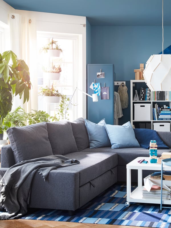 Living room with a corner sofa at centre, surrounded by large, filled shelving units, big windows and many plants.