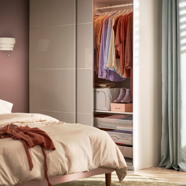 A bedroom with a bed and a PAX/HOKKSUND wardrobe with sliding doors containing clothes hanging on a rod, shoes, and drawers.