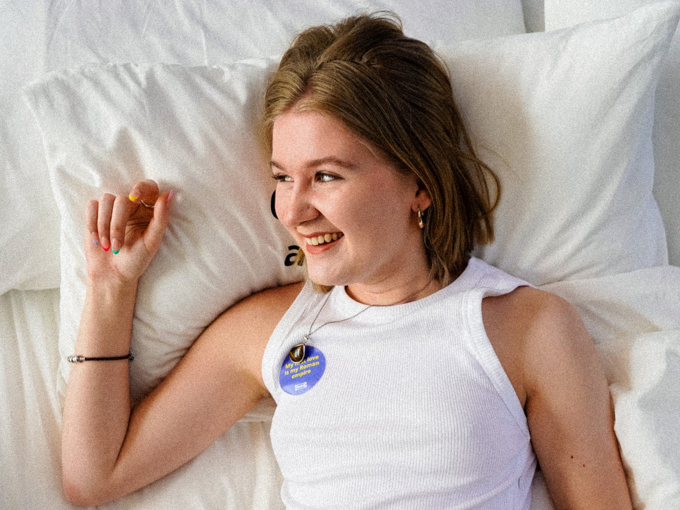 Izzy is lying on a bed in a white t-shirt with her face tilted to the right smiling while holding her right hand up to her face.