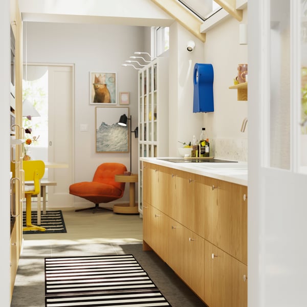 A parallel kitchen with a dining area in the background has SINARP fronts in oak veneer and a striped rug on the floor.