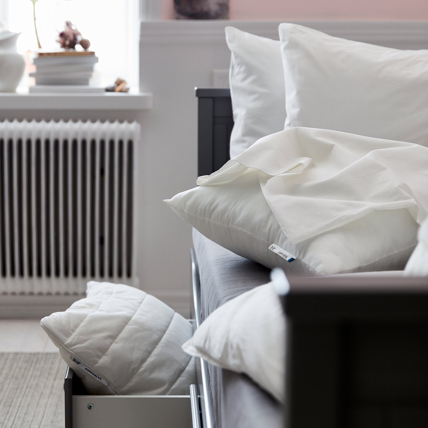 A bed holding a bunch of pillows. The bedframe has a pull-out drawer at the bottom and inside holds another white pillow.