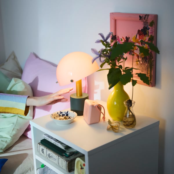 A NÖDMAST LED portable lamp casting a cosy light from a bedside table with pastel decoration next to a bed with pink pillows.