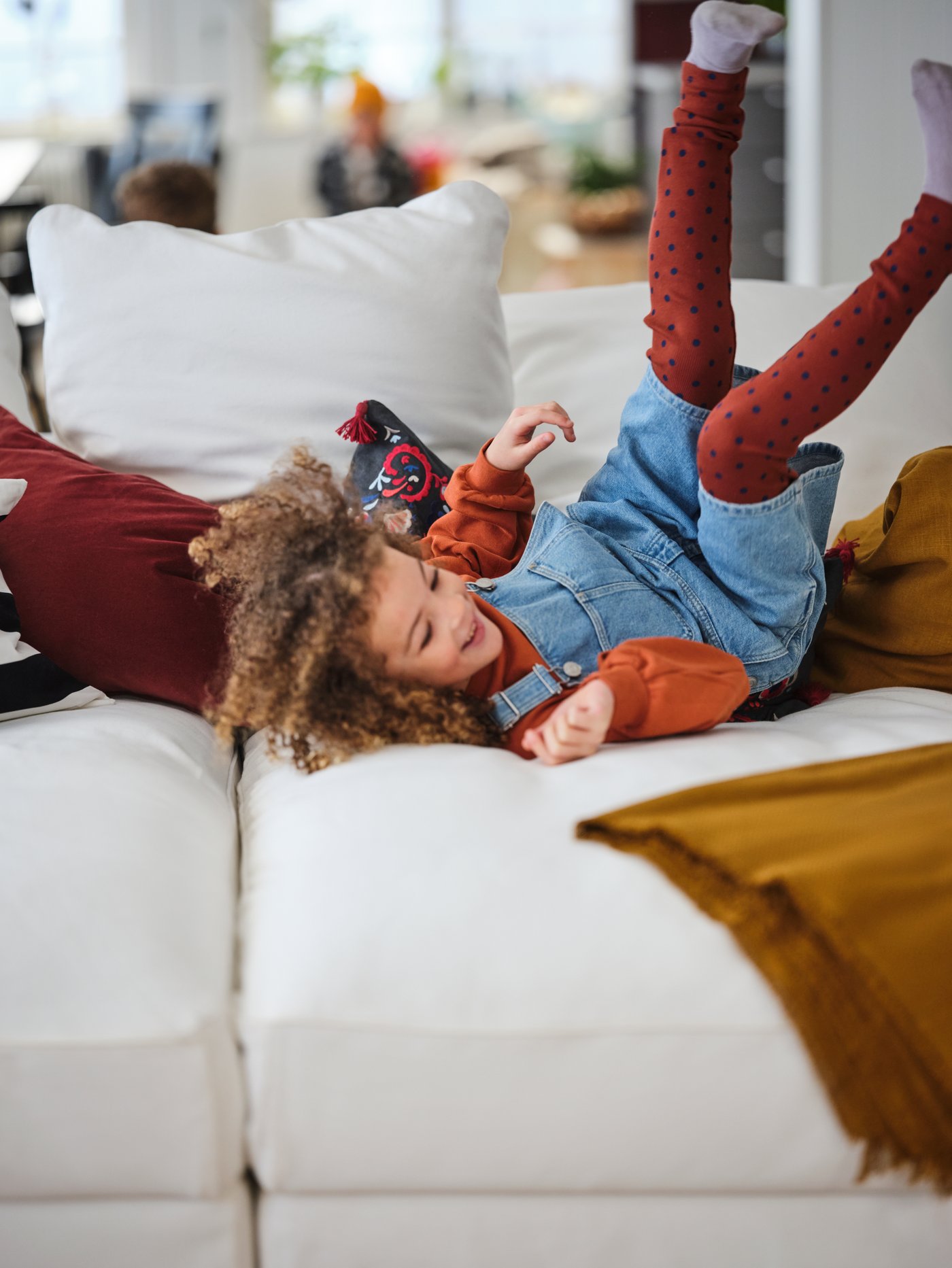 A child happily rolls around, feet in the air, amid colourful textiles on a white GRÖNLID 4-seat sofa with chaise longue.