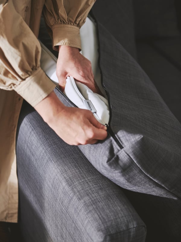 A pair of hands placing a folded-up mattress pad into a dark-grey KLAGSHAMN back cushion on a dark-grey FRIHETEN sofa-bed.