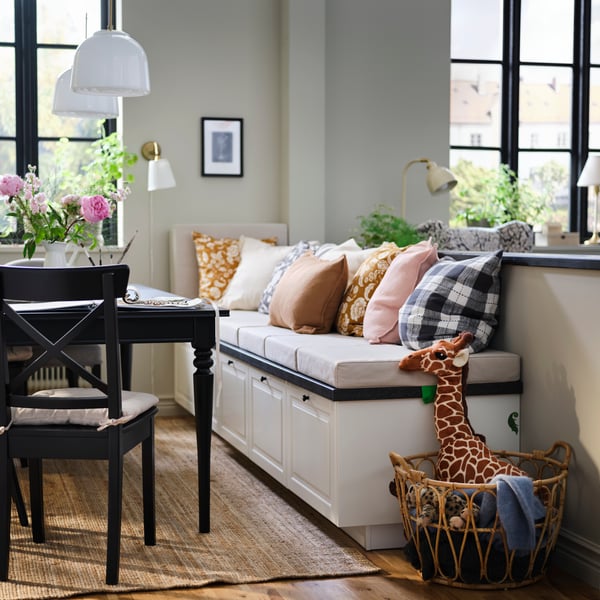 A dining nook with BODBYN drawers in off-white under a bench, decorated with cushions, flowers, and a basket of toys.