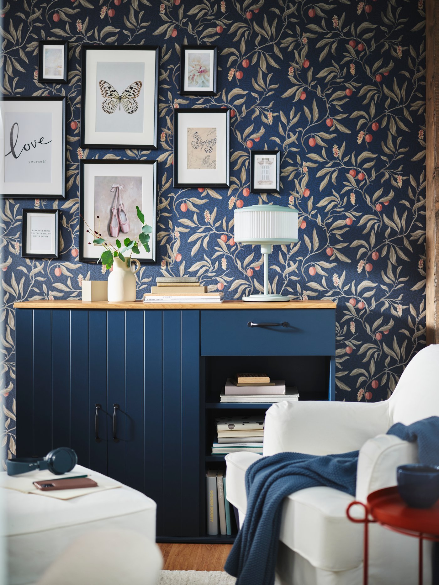 Ein 8-teiliges KNOPPÄNG Set an Rahmen mit Bildern hängt an einer Wand über einem schwarzblauen SKRUVBY Sideboard, vor dem ein ROCKSJÖN Sessel steht.