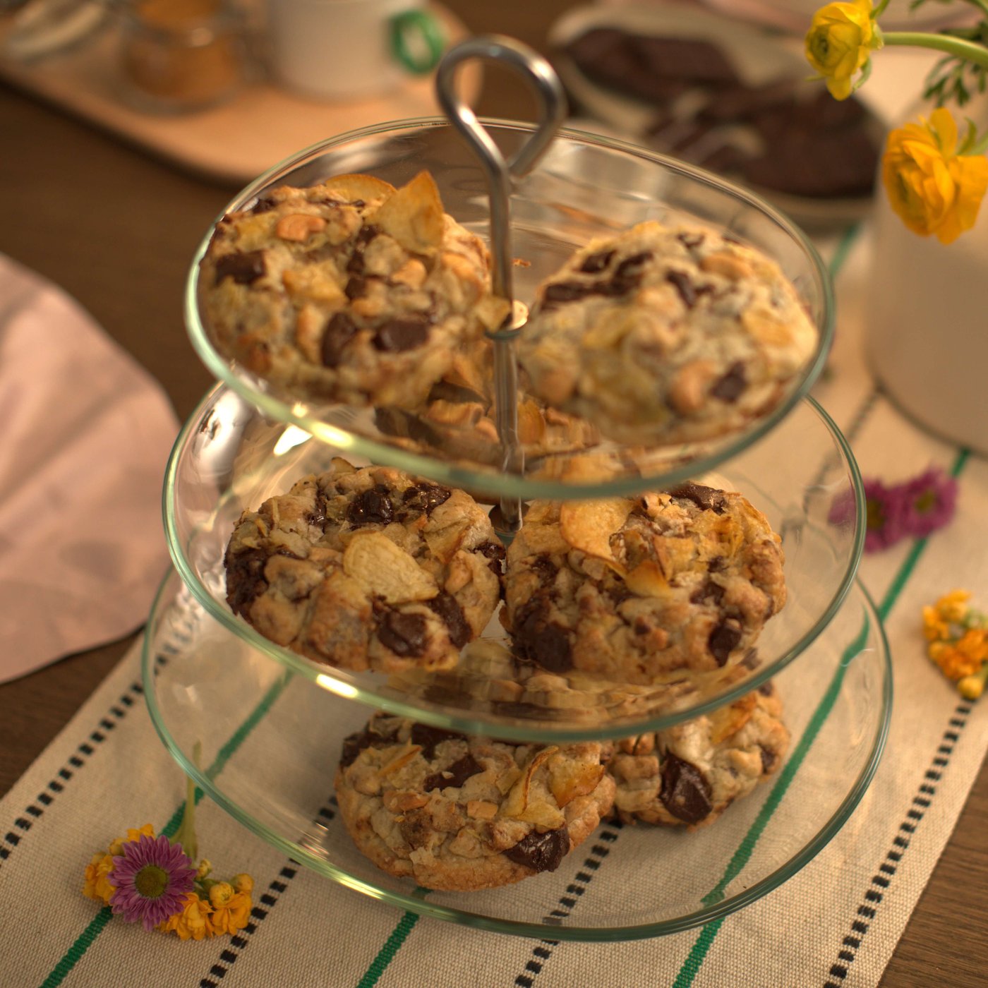Tiered glass stand holding chunky chocolate chip cookies on a table with flowers.