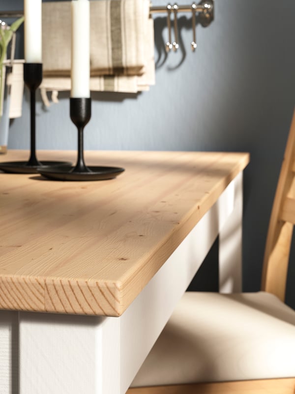 Mesa LERHAMN con cubierta color madera y estructura blanca, con dos candelabros negros en la parte superior, junto a una silla color madera con cojín blanco