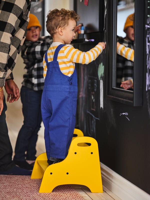 A boy stands on a yellow TROGEN children’s step stool in front of a black TOFTBYN mirror, his brother and father close by.