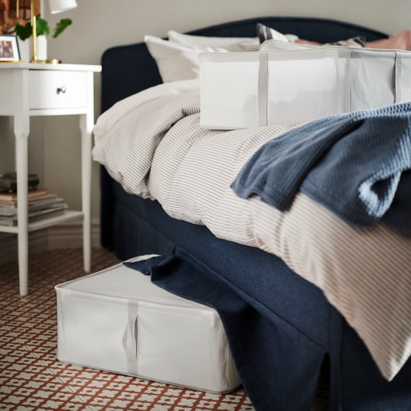 The side of a RAMNEFJÄLL bed with a throw and a HEMMAFIXARE storage case on top. Another storage case is under the bed.