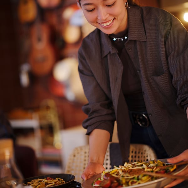 A smiling person in a brown shirt is holding a beige STOCKHOLM 2025 serving bowl with food, about to place it on a table.