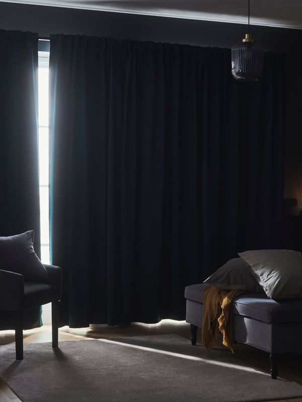 Mörkt rum med gardiner för fönstret, en fåtölj till vänster och en sittpuff med två kuddar och en filt till höger. En taklampa hänger från taket.
