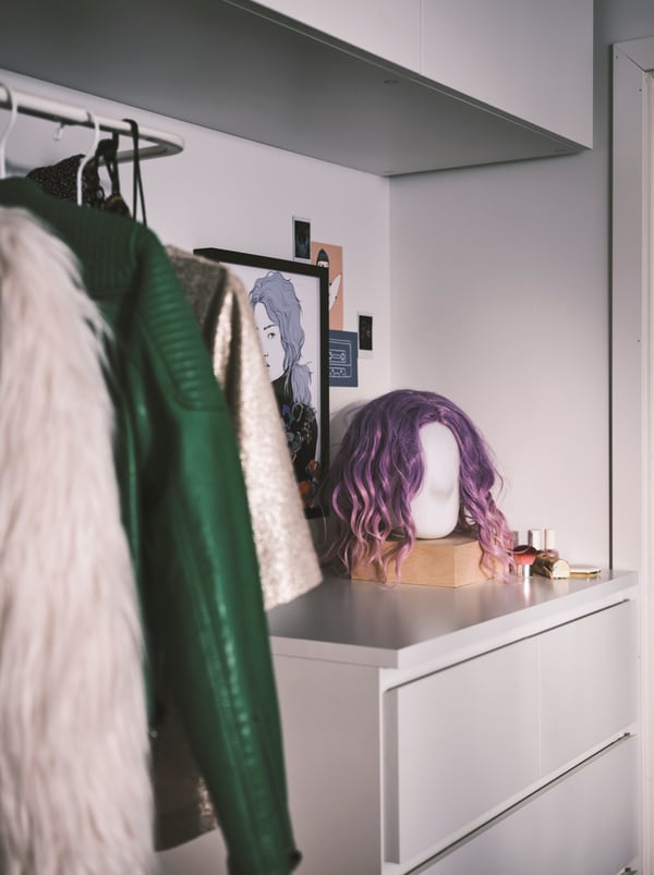 A purple wig on a head shaped lamp that’s on top of a MALM chest with 6 drawers. A hanging rail full of clothes is beside it.