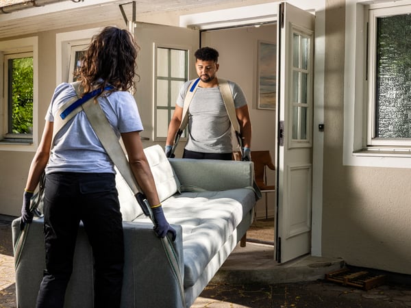 Two people, a woman in the foreground and a man behind her, are carrying a light grey sofa through a doorway using moving straps.