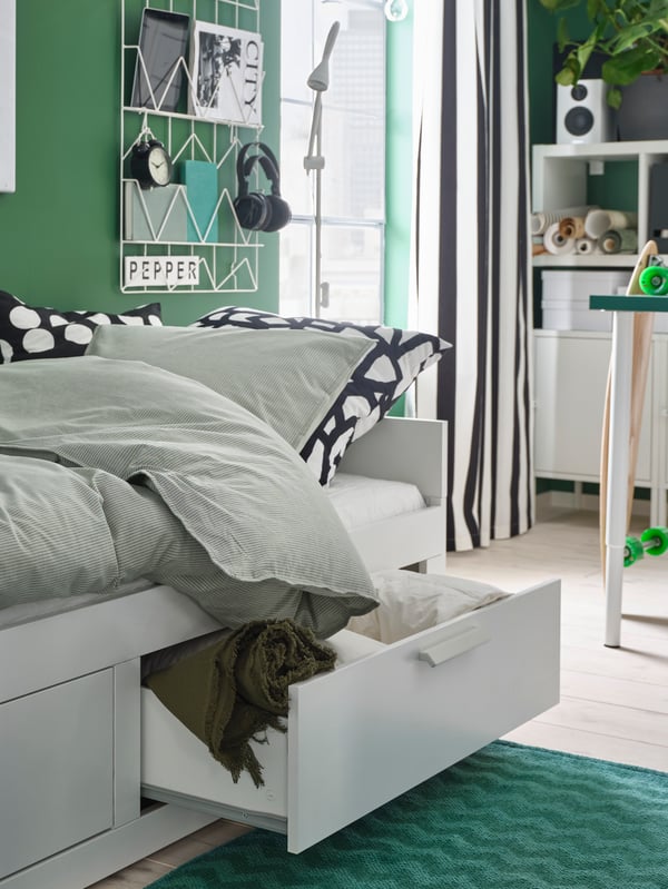 A white BRIMNES day-bed is made as a bed. One drawer is open, and bed linen and a green throw are stored inside.