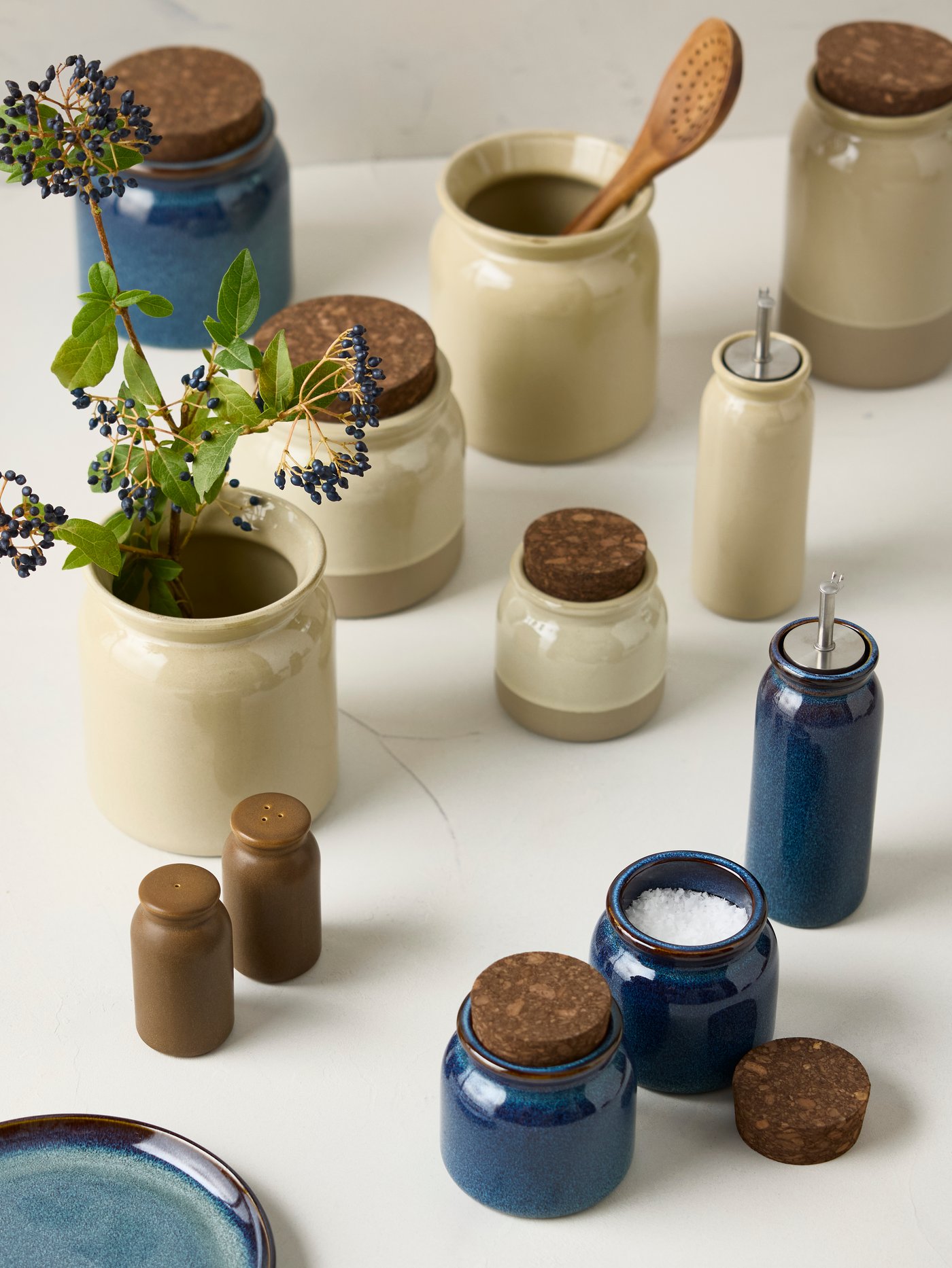 Blue and beige glazed stoneware storage jars some with cork lid, others storing herbs, utensils, olive oil, pepper and salt.