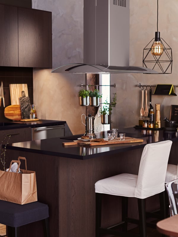 Cocina de madera oscura con una isla que tiene una lámpara de techo con foco BRUNSTA. En la barra hay una cafetera y vasos.