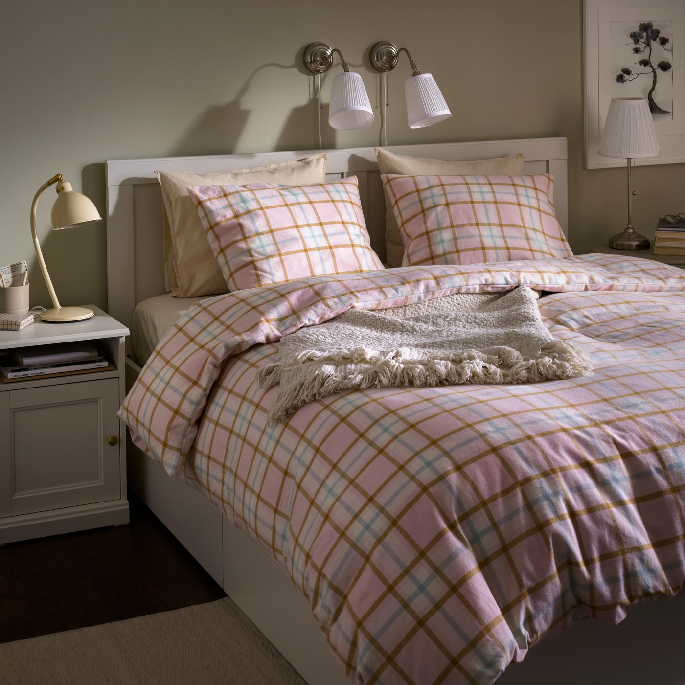 A bedroom with a light pink checked PEROVSKA duvet set on a bed. A white bedside table holds a lamp and books.
