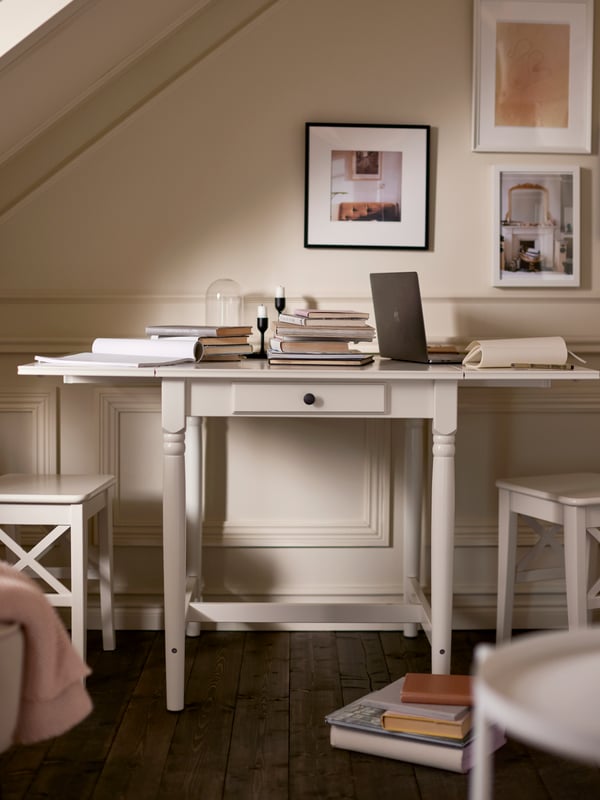 A white INGATORP drop-leaf table with books and a computer on the tabletop, by a bright wall with framed pictures.
