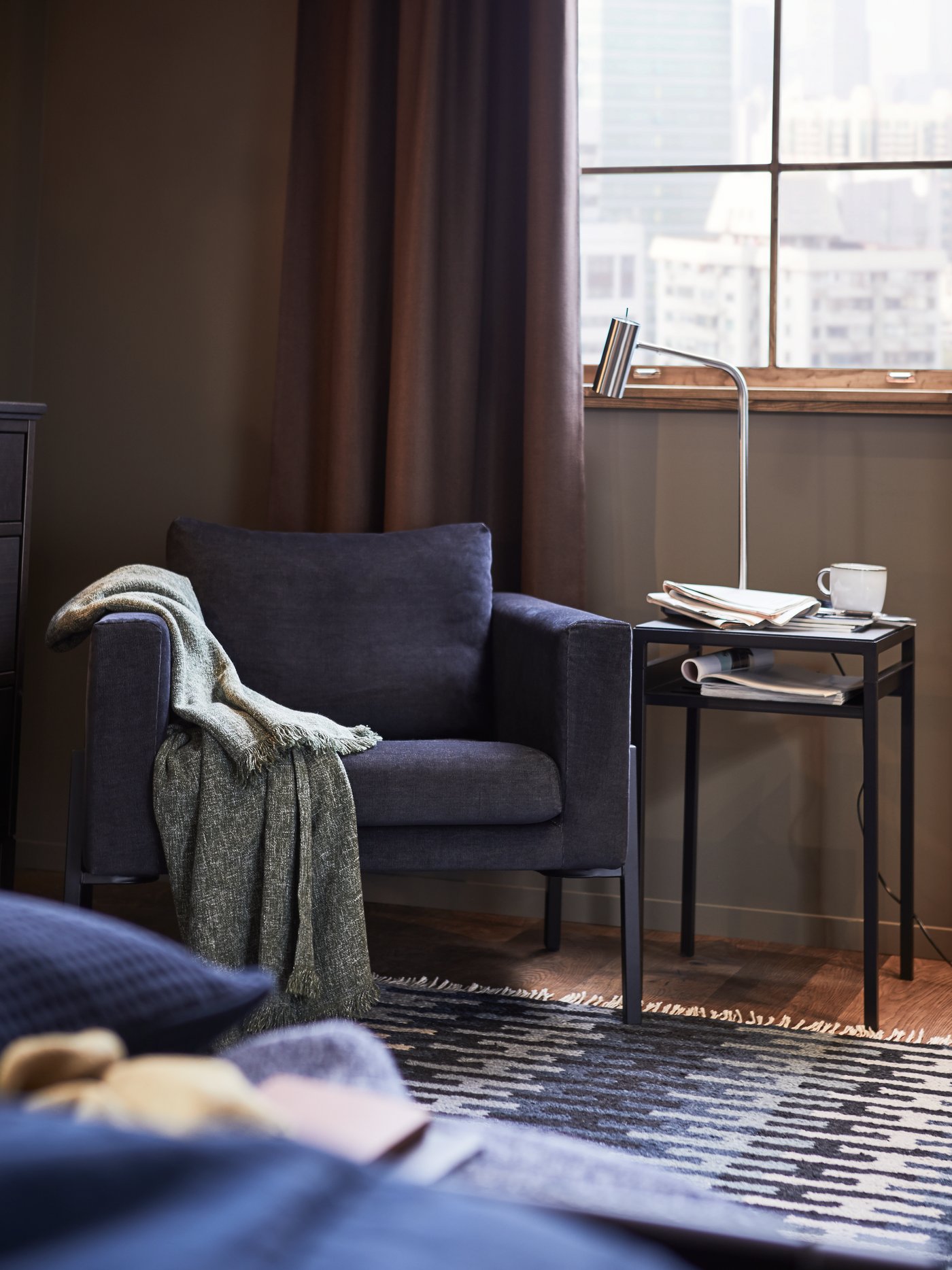 A KOARP armchair with a throw on the right arm, next to a NYBODA side table with a reading lamp, a mug and papers on top.