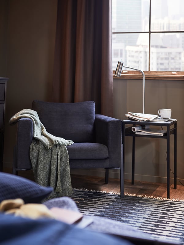 A KOARP armchair with a throw on the right arm, next to a NYBODA side table with a reading lamp, a mug and papers on top.