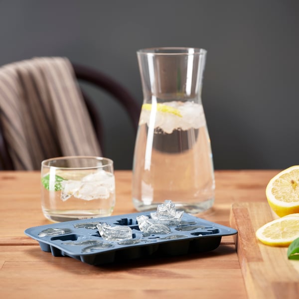 On the table is a dark blue SURSÖT ice cube tray with playful shapes, such as cute ships and whales, next to a carafe and a glass with lemon and mint leaves.