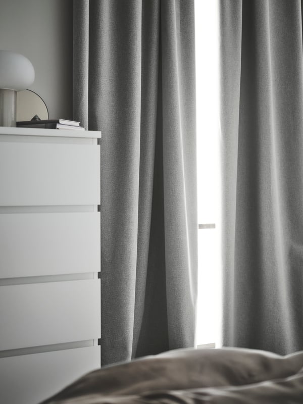 A pair of grey ROSMANDEL room darkening curtains at a window by a white MALM chest of 6 drawers that has a DESJA lamp on it.