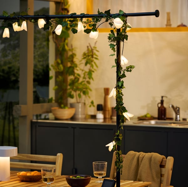 A veranda in evening light. A HELGEÖ decorating rod draped with plants and a glowing lighting chain is attached to a table.