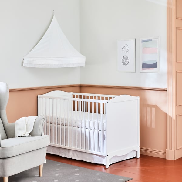 A white SMÅGÖRA cot sits in a corner of a room with a bed canopy on the wall above it and a STRANDMON wing chair beside it.