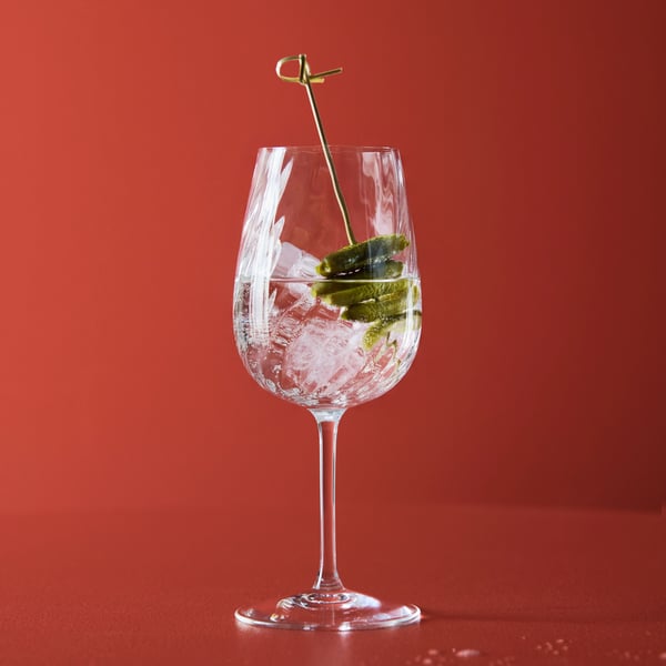 A clear FJÄLLUGGLA glass with an iced sparkling drink and pickled cucumber in it stands against a red background.