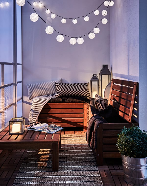 Balkon s dřevěným nábytkem, polštáři, lampami, osvětlením a s květinami. 