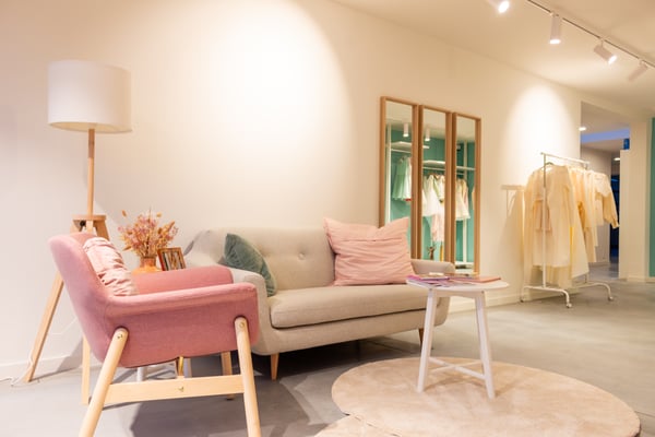 Een gezellige zithoek in een boetiek met pastelgekleurde stoelen, zachte kussens, een kleine tafel, een vloerlamp en kledingrekken op de achtergrond.