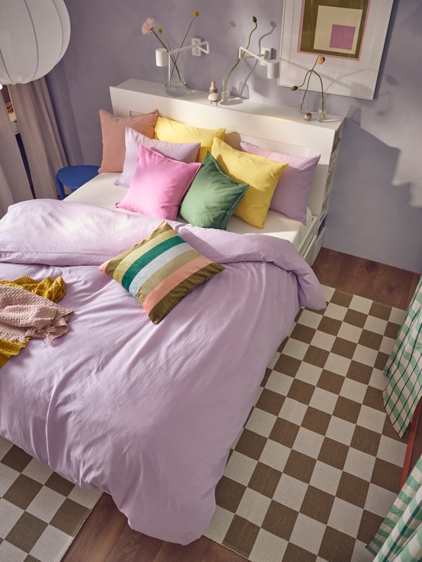 A BRIMNES bed covered with NATTSVÄRMARE bed linen and bright coloured cushions with two KLASSRUM rugs on the floor around it.
