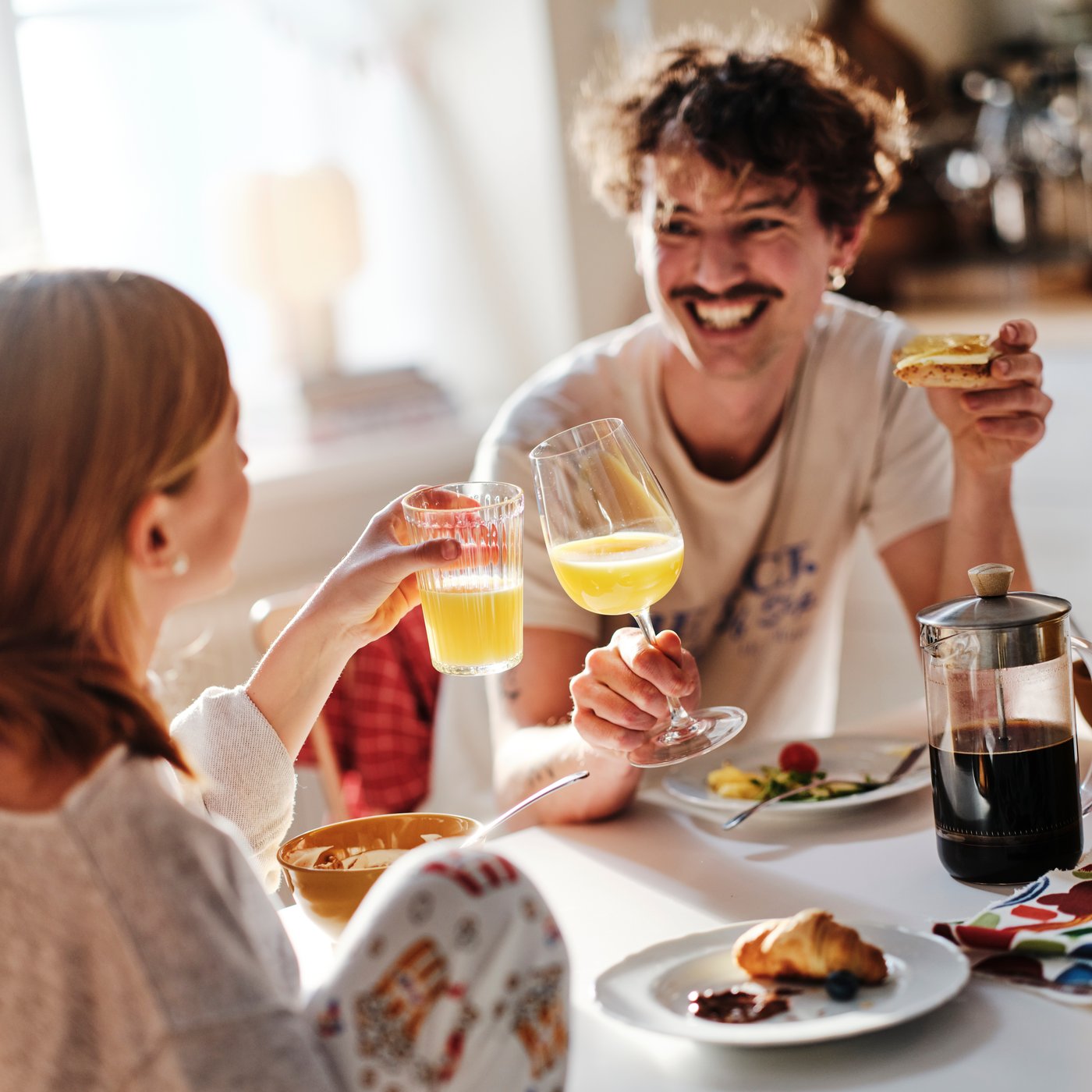 A smiling dad and his teenaged daughter sitting at the breakfast table clink their glasses of orange juice together.