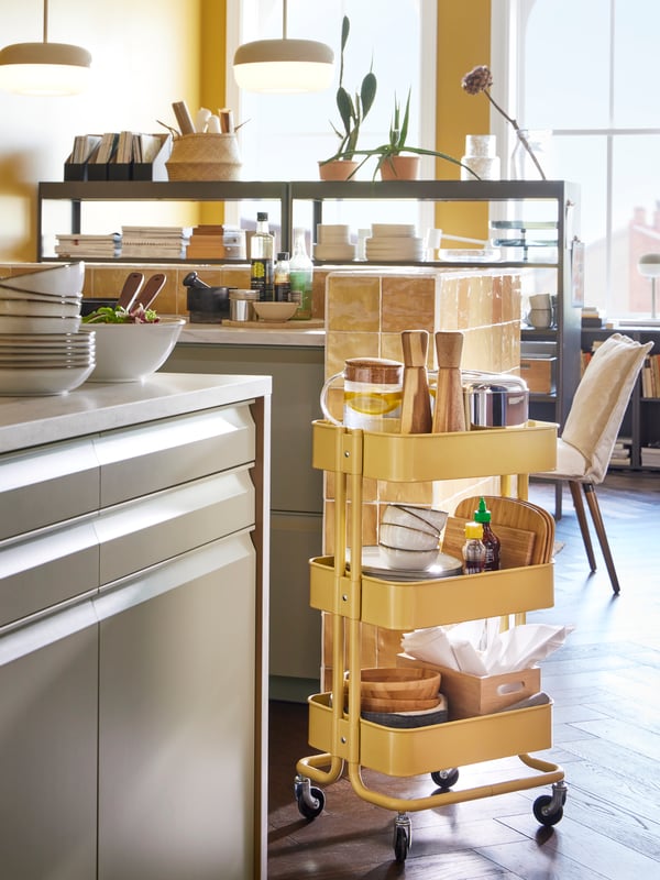 A kitchen with a RÅSKOG trolley in yellow holding diverse items such as bowls, chopping boards, condiments, and a box.