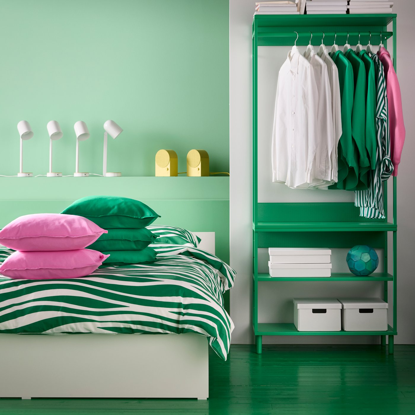 A green PLATSA open shelving unit is placed next to a bed. Bluetooth speakers and desk lamps sit on a niche behind the bed.
