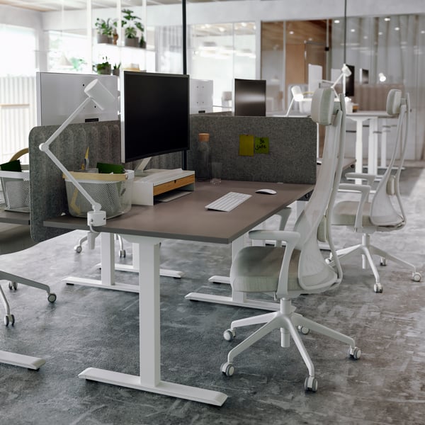Un bureau avec un ordinateur posé sur un bureau assis/debout et un fauteuil blanc poussé en dessous.