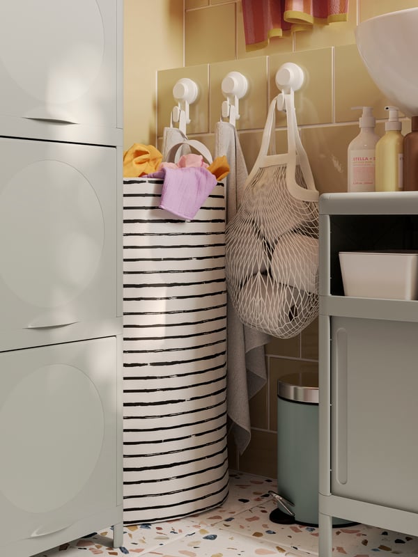A striped KLUNKA laundry bag is standing in a corner and a KUNGSFORS net bag is hanging from a TISKEN hook with suction cup.