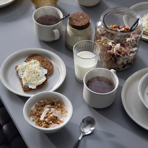 Su un tavolo grigio sono disposte un piatto DAGGASTER con due fette di pane di segale tostato e una ciotola DAGGASTER con muesli e latte.