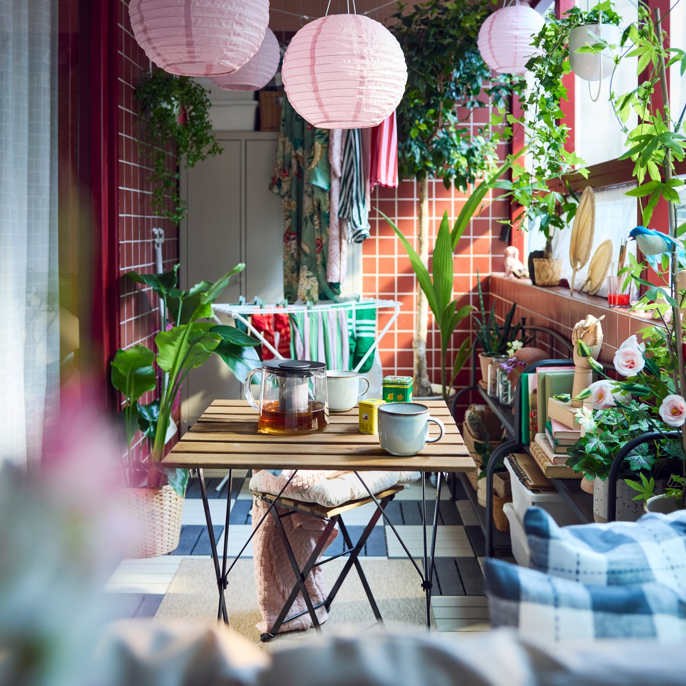 A cosy balcony with green plants and pink lamps, featuring a black and light brown wooden TÄRNÖ outdoor table and tea mugs.