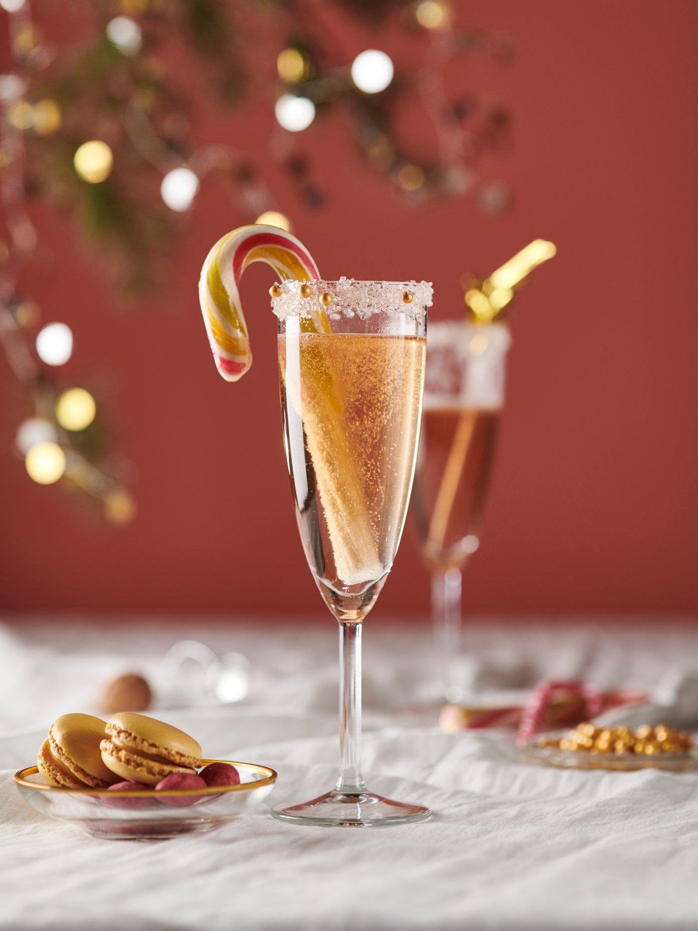 Et STORSINT champagneglas med gylden drik, sukkerkant og rød- og hvidstribet sukkerstang som pynt på et dækket bord med småkager og julelys i baggrunden.